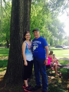 A young woman and elderly man standing with their arm around each other under a tree. 