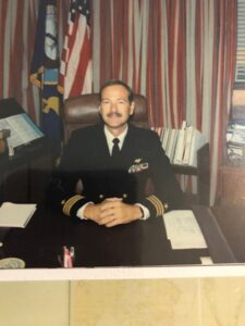 A man in a U.S. Navy uniform sitting behind a desk with clasped hands on the desk.