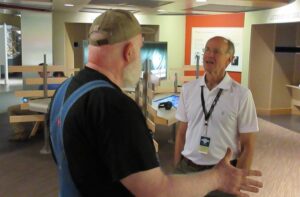 Man in a white shirt with a lanyard talking to another man in overalls and a hat.