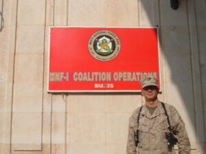 Man in Marine uniform standing under a red sign that reads "MNF-1 Coalition Operations."