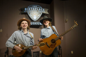 Two boys standing in front of the Birthp[lace of Country Music logo. One is holding a guitar and one holds a mandolin.