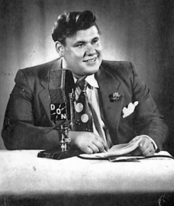 White man in a suit sitting at a desk smiling in front of a microphone